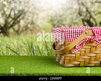 Ein Picknickkorb mit Strohhalm auf einem grünen Rasen vor dem Hintergrund einer atemberaubenden Natur. Camping, Picknick, romantisches Date, leckeres Essen und Getränke. Werbung, Stockfoto