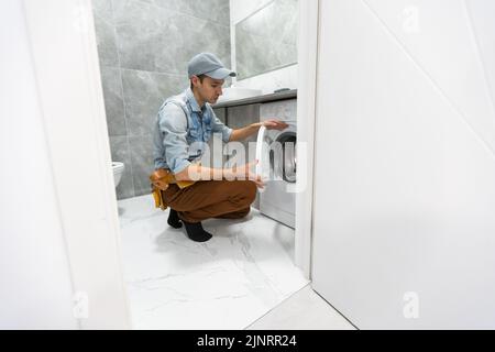 Der junge, gutaussehende Mechaniker im Arbeitsanzug mit der professionellen Werkzeugkiste fixiert die Waschmaschine im Badezimmer. Stockfoto