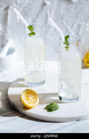 Zwei Gläser eiskalte Limonade mit Minzblättern und Strohhalmen auf Marmortablett bei hellem Sommersonnenlicht und einer in zwei Hälften geschnittenen Zitrone Stockfoto