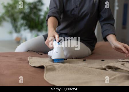 Frau verwendet eine Maschine, um Pellets und Spulen aus Kleidung und Stoff auf schwarzen Hosen zu entfernen. Ein modernes elektronisches Gerät zur Aktualisierung alter Dinge. Stockfoto