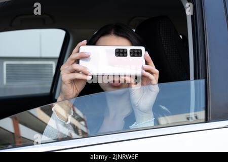 Junge Frau macht Fotos auf dem Handy aus dem Auto Seitenfenster. Viele Kameras auf dem Smartphone. Spionage mit Humor Konzept Stockfoto