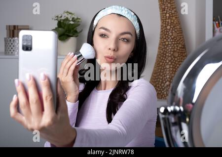 Influencer, Beauty-Blogger, Content-Creator oder Vlogger machen ein Selfie auf einem Handy-Frontkamera. Attraktive junge Frau hält einen kosmetischen Pinsel f Stockfoto