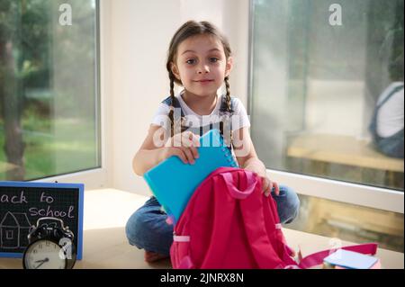 Niedliches kaukasisches Schulmädchen, das auf einem Tisch neben einem Wecker und einem Kreidetafel sitzt, die Kamera anschaut und eine Schultasche zusammenfaltet Stockfoto