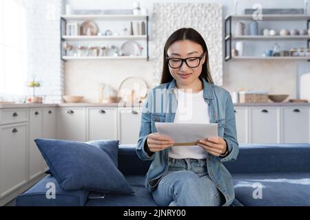 Junge schöne asiatische Studentin erhielt einen glücklichen Brief von der Universität, Teenager-Mädchen erhielt Nachrichten über Studienbeginn und bestanden Prüfungsergebnisse, sitzen auf dem Sofa zu Hause im Wohnzimmer Stockfoto