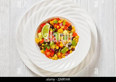 Cowboy-Bohnensalat, texas-Kaviar mit schwarzer Bohne, Tomaten, Avocado, roter Paprika, Mais, Koriander in weißer Schale auf weißem Tisch, flach liegend, cloe-up Stockfoto