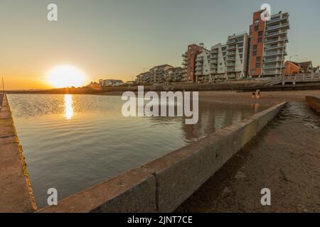 Southend on Sea, Großbritannien. August 2022. Eine ruhige Küstenszene bei Sonnenuntergang mit einem Gezeitenbecken oder einer Meeresmauer im Vordergrund, mit goldenem Licht, das vom Wasser reflektiert wird. Moderne Wohnhäuser säumen die Uferpromenade im Hintergrund und veranschaulichen den Kontrast zwischen natürlicher Schönheit und städtischer Entwicklung. Sonnenuntergang am Chalkwell Beach, in der Nähe von Southend on Sea. Bei Temperaturen um 25 Grad nutzen viele Leute den kühleren Abend. Penelope Barritt/Alamy Live News Stockfoto