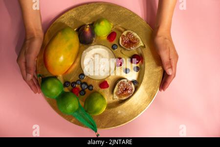 Die Hände der Frau servieren einen wunderschön servierten Teller mit frischen tropischen Früchten auf einer rosafarbenen Tischansicht. Gesichtsloses Mädchen, das auf dem Tisch serviert wird. Feigen, ma Stockfoto