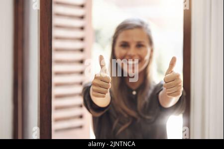 Daumen nach oben von einer glücklichen Frau lächelnd, während eine Hand gewinnende Geste in einem modernen Büro zeigt. Fröhliche, positive Frau, die nach dem Erhalten aufgeregt aussieht Stockfoto