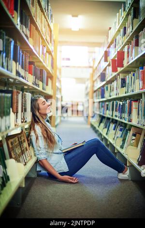 Das Leben im College kann manchmal ziemlich ermüdend sein. Ein Universitätsstudent schläft zwischen den Bücherregalen in der Bibliothek auf dem Campus. Stockfoto
