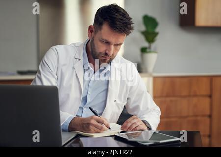 Ein hübscher männlicher Arzt, der in seinem Büro sitzt und in sein Tagebuch schreibt. Stockfoto