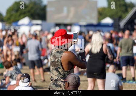 Ein schwarzer Mann mit rotem Eimer Hut und weißer Sonnenbrille hält ein Handy in der Luft, um ein Video auf einem multikulturellen Outdoor-Festival zu machen Stockfoto