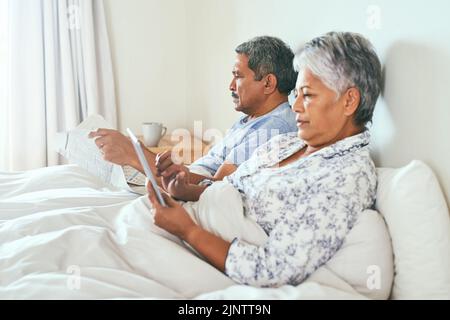 Die einfachen Dinge im Leben sind immer großartig. Ein entspanntes reifes Paar liegt im Bett, während es auf einem digitalen Tablet stöbert und zu Hause die Zeitung liest Stockfoto