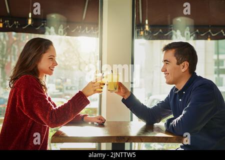 Ein junger Mann und eine junge Frau, die bei einem romantischen Date in einem Café mit ihren Getränken toasten. Stockfoto
