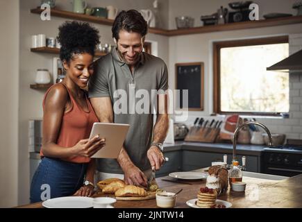 Junges Paar kocht gemeinsam gesundes Essen nach Rezepten online auf einer Tablette, Schritt für Schritt. Glücklich, fröhlich und lächelnd Mann und Frau machen Stockfoto
