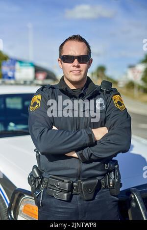 Ich bin bei der Arbeit. Ein beschnittenes Porträt eines hübschen jungen Polizisten, der mit gekreuzten Armen auf der Patrouille steht. Stockfoto