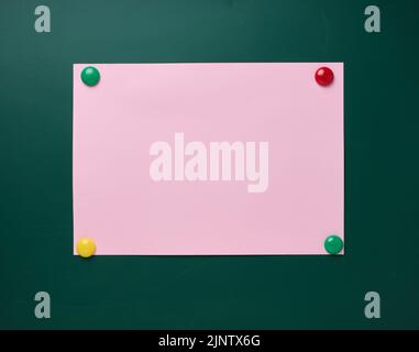 Ein leeres rosafarbenes Blatt wird mit Magneten an einer grünen Schultafel befestigt, ein Platz für eine Beschriftung Stockfoto