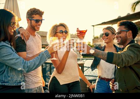 Mit Freunden wie diesen gibt es viel zu feiern. Eine Gruppe junger, vielfältiger Freunde, die einen Drink genießen und den Tag draußen auf dem Dach verbringen. Stockfoto