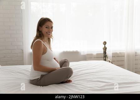 Attraktive Schwangeren sitzt auf dem Bett Lächeln schaut auf die Kamera Stockfoto