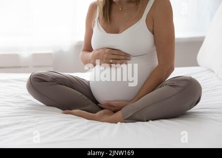 Schwanger Frau sitzt mit gekreuzten Beinen auf dem Bett berühren Bauch Genießen Sie die Schwangerschaft Stockfoto