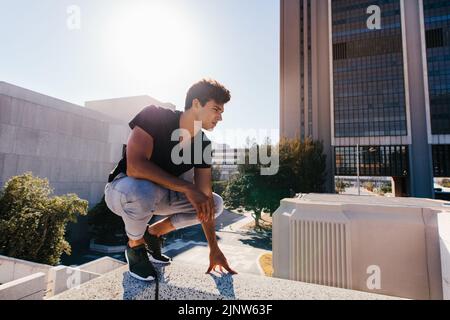 Junger Sportler hockt im Freien, bevor er Parkour und Freilauftechniken vorführt. Fit junger Mann immer bereit, um in CI zu üben Stockfoto