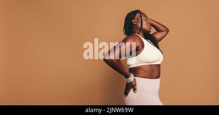 Sportliche Frau, die mit geschlossenen Augen vor einem braunen Hintergrund steht. Reife Frau mit Dreadlocks in Sportkleidung in einem Studio. Sportlicher Blac Stockfoto