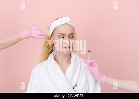 Die Frau wählt auf beiden Seiten ein Gesichtspflegeverfahren, die Hände mit Handschuhen reichen in einer Spritze im zweiten Pinsel zur Anwendung therapeutischer Masken zu ihr aus. In einem weißen Mantel an einer rosafarbenen Wand stehend Stockfoto