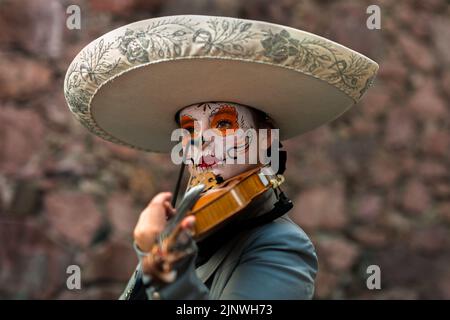 Ein mexikanisches Mädchen, bekleidet als La Catrina und mit einem Mariachi Sombrero Hut, spielt die Geige während der Day of the Dead Feierlichkeiten in Taxco, Mexiko. Stockfoto