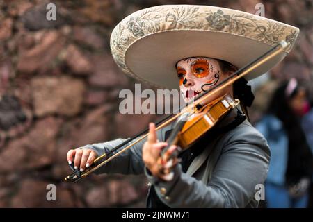 Ein mexikanisches Mädchen, bekleidet als La Catrina und mit einem Mariachi Sombrero Hut, spielt die Geige während der Day of the Dead Feierlichkeiten in Taxco, Mexiko. Stockfoto