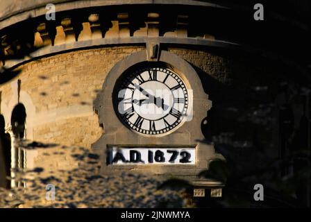 Zeit zum Nachdenken: Das Baudatum 1872 auf dem Uhrturm der ehemaligen Bliss Tweed Mill in Chipping Norton, Oxfordshire, England, Großbritannien, spiegelt sich in der ruhigen Oberfläche des kleinen Sees in Landschaftsgärten wider, die von den heutigen Bewohnern, den Pächtern von Luxuswohnungen, die nach der Schließung der Mühle im Jahr 1980 entstanden, genossen wurden. Stockfoto