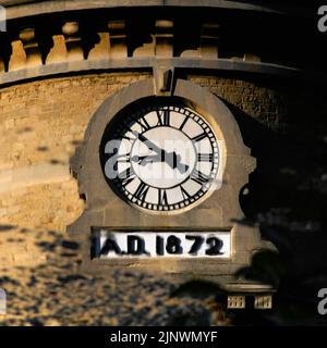 Quadratische Zeiten: Das Baudatum 1872 auf dem Uhrturm der ehemaligen Bliss Tweed Mill in Chipping Norton, Oxfordshire, England, Großbritannien, spiegelt sich in der ruhigen Oberfläche des kleinen Sees in Landschaftsgärten wider, die von den heutigen Bewohnern, den Pächtern von Luxuswohnungen, die nach der Schließung der Mühle im Jahr 1980 entstanden, genossen wurden. Stockfoto