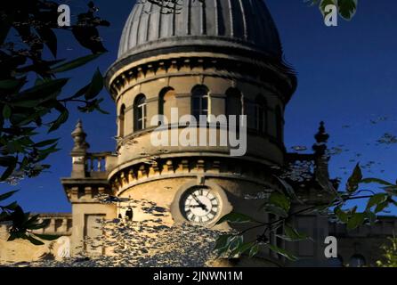 Die Zeit vergeht und die Industriezeit vergeht: Das Baudatum 1872 auf dem Uhrenturm der ehemaligen Bliss Tweed Mill in Chipping Norton, Oxfordshire, England, Großbritannien, spiegelt sich in diesem quadratischen Format in der ruhigen Oberfläche des Sees in den Landschaftsgärten wider, die von den heutigen Bewohnern genossen werden. Die Pächter von Luxuswohnungen, die nach der Schließung der Mühle im Jahr 1980 entstanden sind. Stockfoto