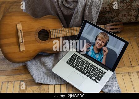 Süßer blonder Junge mit Kopfhörern spielt Gitarre und schaut auf den Laptop. Eine lustige Online-Lektion im Internet. Digitale Bildung. Moderner Lifestyle in Stockfoto