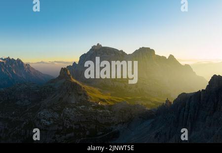 Blick von oben, atemberaubende Luftaufnahme einer Bergkette bei einem schönen Sonnenaufgang mit einer Berghütte in der Ferne. Stockfoto