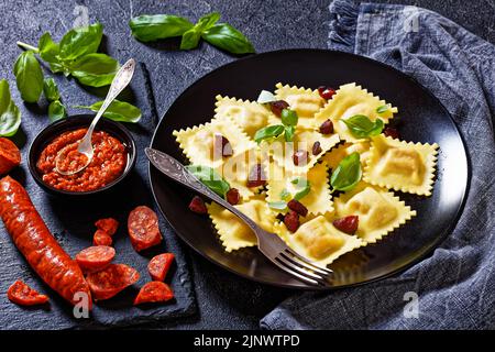 italienische Ravioli mit Chorizo Ricotta Füllung auf schwarzem Teller mit gebratenem Chorizo serviert, frisches Basilikum auf dunklem Betontisch mit Zutaten, horizontal Stockfoto