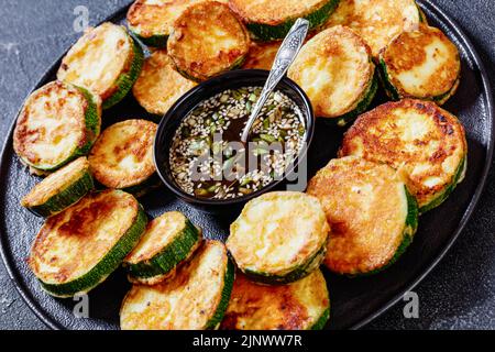 Nahaufnahme von Hobak Jeon, in der Pfanne gebratene Zucchini in Eierteig serviert mit Soja-Dipping-Sauce auf schwarzem Teller auf Betontisch, koreanische Küche Stockfoto