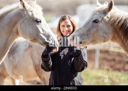 Junge Frau in schwarzer Reitjacke in der Nähe einer Gruppe von weißen arabischen Pferden, die glücklich lächeln, einer auf jeder Seite, Nahaufnahme Detail Stockfoto