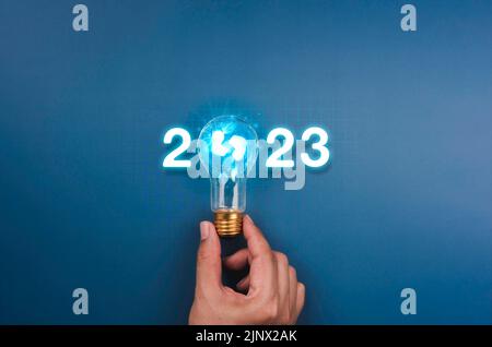 Leuchtende Kalenderjahreszahlen 2023, Neon-Stil mit kreativer Trend-Glühbirne, die von Geschäftsleuten auf digitalem Netzwerk gehalten wird, und blauer Hintergrund. Ha Stockfoto