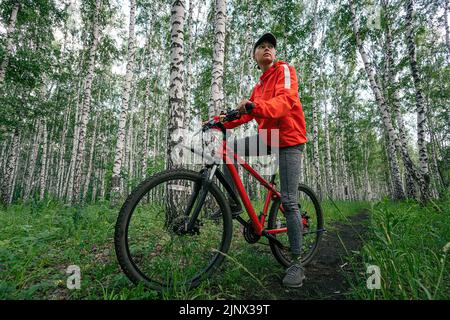 Eine Radfahrerin fährt auf der Straße in einem wunderschönen dichten grünen Wald mit dem Rennrad. Bikegirl in Mystery Forest. Stockfoto