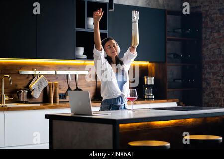 Glückliches Mädchen mit prothetischen Arm Musik hören Kopfhörer Küche zu Hause, Frau mit Behinderung entspannend und tanzen. Stockfoto