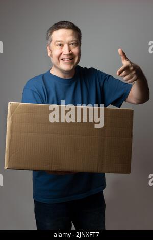 Kaukasischer Mann mit einem Lächeln, das auf die große Lieferkiste zeigt, die er in der Hand hält Stockfoto