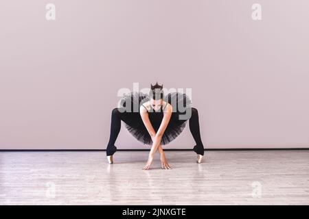 Anmutige Ballerina in schwarzem Schwanenkleid vor wgite Hintergrund. Junge Balletttänzerin, die vor der Aufführung in schwarzem Tutu, klassischem Tanzstud, praktiziert Stockfoto