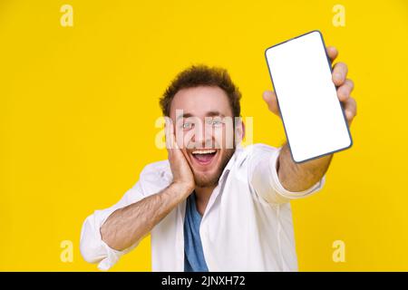 Tolles Angebot. Junger glücklicher Mann hält Smartphone zeigt einen weißen leeren Bildschirm und spannend zu gewinnen isoliert auf gelbem Hintergrund, feiert Erfolg. Produktplatzierung. Werbung Für Mobile Apps. Stockfoto