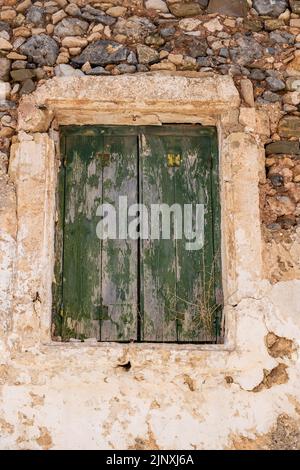 Veraltete hölzerne geschlossene Fensterläden, geschälte Steinmauer griechische ländliche Gebäudefassade. Verlassene Haus Hintergrund, grün bemalte Fensterbretter. Vertikal Stockfoto