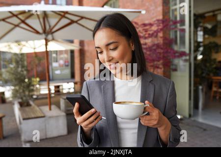 Junge elegante asiatische Frau hält Kaffee im Freien stehend mit dem Smartphone. Stockfoto