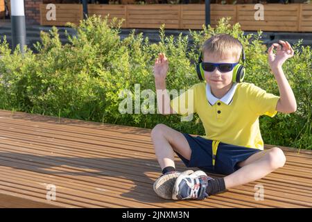 Ein stilvoller Junge mit Sonnenbrille und Kopfhörern hört seine Lieblingsmusik, während er auf einer Bank in einem Stadtpark sitzt. Ein glücklich lächelnder kleiner Junge genießt m Stockfoto