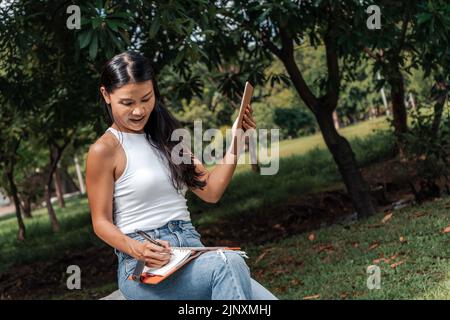 Glückliche Frau, die in einem Park ein Notizbuch schreibt und benutzt Stockfoto