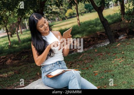 Glückliche Frau, die in einem Park ein Notizbuch schreibt und benutzt Stockfoto