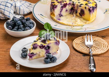 Heidelbeer-Joghurt-Kuchen in weißer Keramikschale. Portionieren Sie Heidelbeer-Joghurt-Kuchen mit Heidelbeeren in Keramikbecher, Metallgabel auf Bambusmatte auf Holztisch. Stockfoto