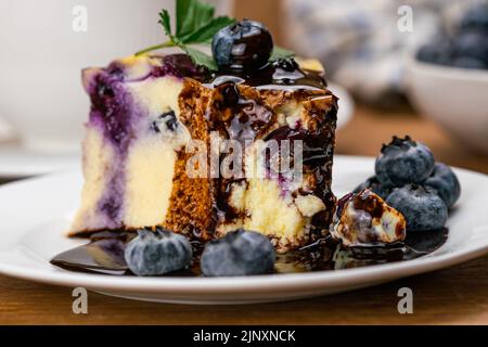 Extreme Nahaufnahme von Heidelbeer-Joghurt-Kuchen mit Schokoladensirup in weißer Keramikschale mit einer Tasse Heidelbeeren-Obst auf Holztisch gegossen. Stockfoto