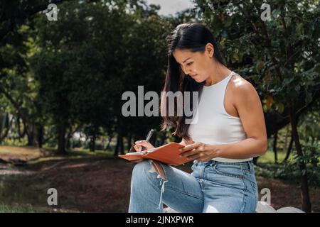Glückliche Frau, die in einem Lehrbuch in einem Park schreibt Stockfoto